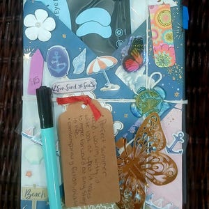 May include: A gift package wrapped in patterned paper featuring beach-themed stickers, a black eye mask packet, a light blue pen, a brown paper tag with handwritten text, a gold butterfly cutout, and a wax seal, all tied together with white string.