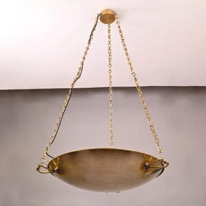 May include: A brass pendant light fixture featuring a wide, shallow bowl-shaped shade suspended by three decorative metal chains that connect to a central circular ceiling canopy. The metal has a warm, polished finish with visible texture.