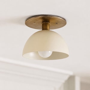 May include: A ceiling light with a cream-coloured, dome-shaped shade and a brass-coloured base. A light bulb is visible inside the shade. The fixture is mounted on a white ceiling.