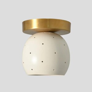 May include: Cream-colored, spherical ceiling light fixture with small, evenly spaced black dots. The fixture has a flat, gold-colored disc on top. The design is modern and minimalist, suitable for various interior styles.
