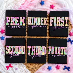 May include: Six black t-shirts with school grade level text in colorful patchwork letters. The shirts read "PREK", "KINDER", "FIRST", "SECOND", "THIRD", and "FOURTH" grade. Decorative stars and bows surround the shirts.