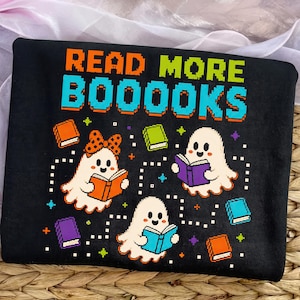 May include: A black t-shirt featuring a pixel art graphic with the text READ MORE BOOOOKS in orange and blue. The design includes three white ghosts reading colourful books, with small scattered pixelated stars and squares in various colours.