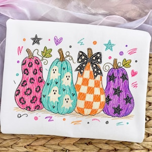 May include: A white sweatshirt featuring a colorful embroidered design of four pumpkins. The first is pink with black leopard spots, the second is teal with white ghosts, the third is orange with a checkered pattern and a black bow, and the fourth is purple with black stars.