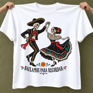 Peut inclure: Un t-shirt blanc à manches courtes avec un motif de deux squelettes dansant en tenue mexicaine traditionnelle. Le squelette masculin porte un costume de charro et un sombrero, tandis que la femelle porte une robe à volants. Texte : BAILAMOS PARA RECORDAR.
