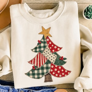 Könnte beinhalten: Ein cremefarbenes Sweatshirt mit Rundhalsausschnitt und einer großen, mittigen Grafik eines Weihnachtsbaums im Patchwork-Stil. Der Baum besteht aus verschiedenen Stoffmustern, darunter grün-weißes Vichy-Karo, rot-weiße Streifen und rote Punkte.