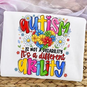 May include: White t-shirt with a colorful graphic design. The design includes the word "Autism" in rainbow colors above a heart-shaped arrangement of flowers. The text "IS NOT A DISABILITY It's a different ABILITY" is also displayed.