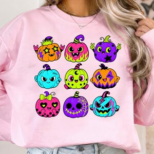 Puede incluir: Una sudadera rosa claro con un estampado gr&aacute;fico de nueve calabazas de dibujos animados coloridas. Las calabazas est&aacute;n decoradas con varios patrones, incluyendo estrellas, rayos, llamas, lunares y telara&ntilde;as en colores ne&oacute;n.