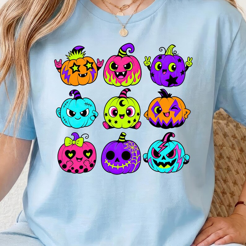 Puede incluir: Una camiseta azul claro con un estampado gr&aacute;fico de nueve calabazas de dibujos animados coloridas. Las calabazas muestran diversas expresiones y patrones, incluyendo estrellas, rayos, lunares y telara&ntilde;as, en tonos naranja, rosa, morado y turquesa.