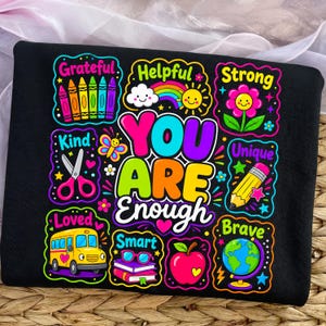 May include: A black t-shirt featuring a colorful graphic with the text "YOU ARE Enough" surrounded by illustrations of crayons, a rainbow, a sun, a flower, scissors, a butterfly, a pencil, a school bus, glasses, books, an apple, and a globe.