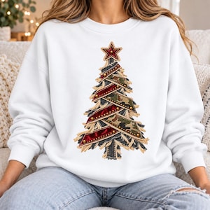 May include: A white crewneck sweatshirt featuring a central graphic of a stylised Christmas tree. The tree is constructed from layered strips of red velvet, camouflage fabric, and frayed denim, accented with small gold studs and a red star.