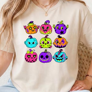 Puede incluir: Una camiseta beige de manga corta con un estampado gr&aacute;fico de nueve calabazas de dibujos animados coloridas. Las calabazas est&aacute;n decoradas con varios patrones que incluyen estrellas, llamas, lunares y telara&ntilde;as en tonos ne&oacute;n vibrantes de rosa, naranja, azul y morado.