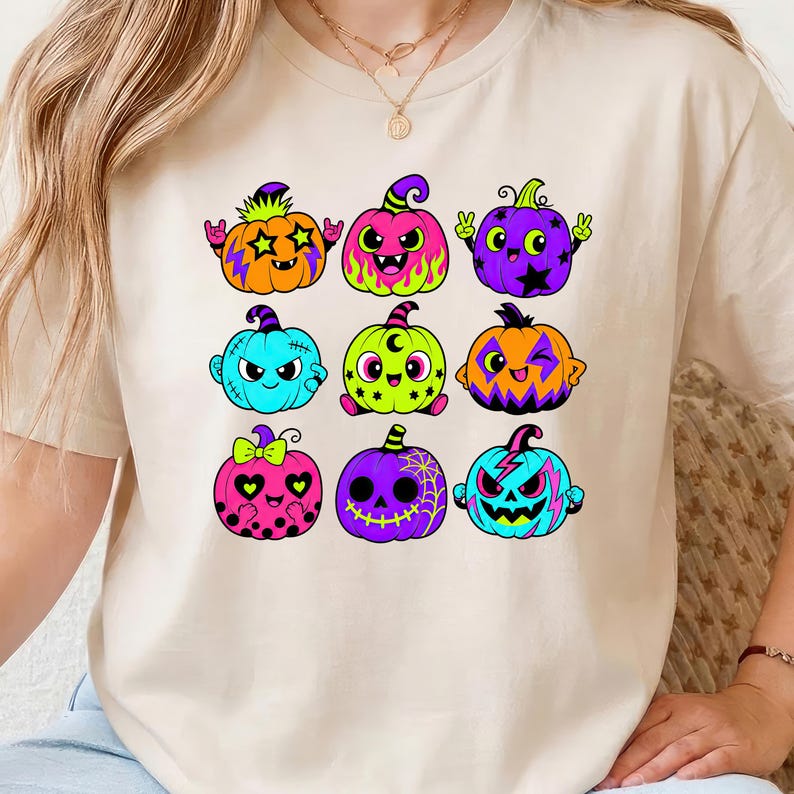 Puede incluir: Una camiseta beige de manga corta con un estampado gr&aacute;fico de nueve calabazas de dibujos animados coloridas. Las calabazas est&aacute;n decoradas con varios patrones que incluyen estrellas, llamas, lunares y telara&ntilde;as en tonos ne&oacute;n vibrantes de rosa, naranja, azul y morado.