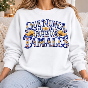 Peut inclure: Un sweat-shirt blanc à col rond arborant un motif graphique avec le texte "QUE NUNCA FALTEN LOS TAMALES" en lettres bleues stylisées. Le design comprend des accents floraux jaunes et orange ainsi que deux tamales enveloppés dans des feuilles.