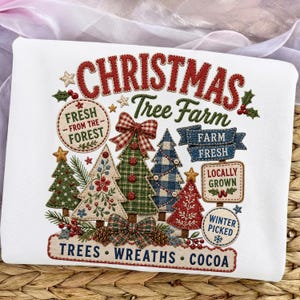 May include: A white sweatshirt featuring a colorful embroidered design with the text CHRISTMAS Tree Farm, FRESH FROM THE FOREST, FARM FRESH, LOCALLY GROWN, WINTER PICKED, and TREES WREATHS COCOA. The graphic includes various stylized Christmas trees.