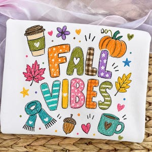 May include: A white sweatshirt featuring a colourful graphic with the text FALL VIBES. The design includes a coffee cup, pumpkin, maple leaf, scarf, acorn, and mug, decorated with hearts, stars, and flowers in orange, green, pink, and blue.