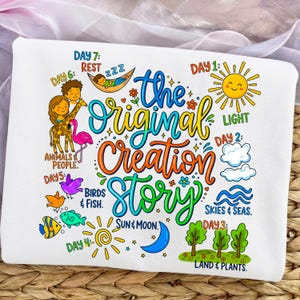 May include: White t-shirt with a colorful graphic illustrating the creation story. The design includes a smiling sun, clouds, trees, and animals, with text such as "Light," "Skies & Seas," and "The Original Creation Story."