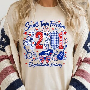 Może przedstawiać: Kremowy t-shirt z grafiką zawierającą napis Small Town Freedom, liczbę 42701 oraz Elizabethtown, Kentucky. Wzór zawiera czerwono-biało-niebieskie motywy, banjo, gitarę, sylwetkę konia oraz kokardę.