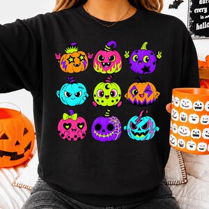 Puede incluir: Una sudadera negra de cuello redondo con un estampado gr&aacute;fico de nueve calabazas de dibujos animados coloridas. Cada calabaza muestra una expresi&oacute;n &uacute;nica y una paleta de colores vibrantes, incluyendo tonos naranja, rosa, morado, verde lima y azul brillante.