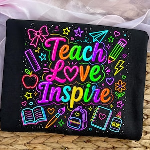 May include: A black t-shirt featuring a vibrant neon-colored graphic with the text Teach Love Inspire. The design includes illustrations of a bow, paper airplane, pencil, apple, ruler, book, paintbrush, backpack, notebook, glue, and various stars and hearts.