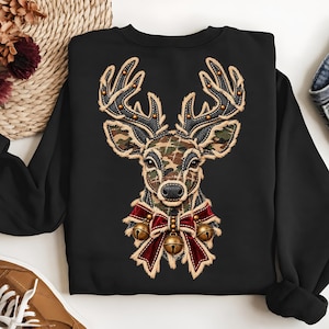May include: A black sweatshirt featuring a central graphic of a deer head with camouflage-patterned fur, large antlers, and a red velvet bow around its neck adorned with three gold-colored jingle bells, all outlined with a frayed fabric effect.
