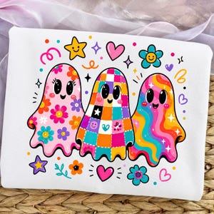 May include: A white sweatshirt featuring a colorful graphic of three ghosts. The left ghost is pink with floral patterns, the middle ghost has a multicolored checkered design, and the right ghost features wavy rainbow stripes. Surrounded by stars, hearts, and flowers.