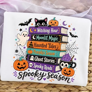 May include: A white sweatshirt featuring a colorful graphic of a stack of books with titles Witching Hour, Moonlit Magic, Haunted Tales, Pumpkin Season, Ghost Stories, and Spooky Reads, surrounded by pumpkins, a ghost, a bat, and a skull mug.
