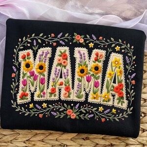 Puede incluir: Sudadera negra con la palabra "MAMA" bordada en un diseño floral. Las letras están rellenas de girasoles, tulipanes y otras flores. Un borde floral rodea las letras, con pequeñas flores y hojas.