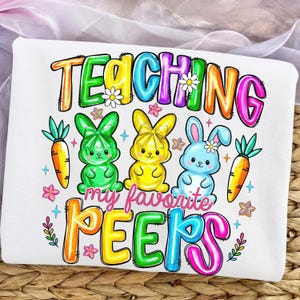 May include: White t-shirt with a colorful graphic featuring the text "TEACHING my favorite PEEPS" above three cartoon bunnies and two carrots. The bunnies are green, yellow, and blue, with flower accents. The text is outlined in various colors.