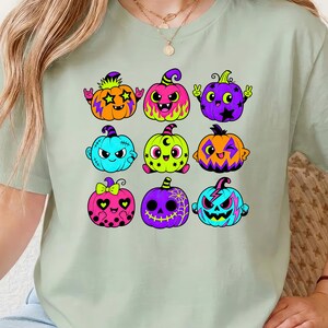 Puede incluir: Una camiseta de color verde salvia claro con un estampado gr&aacute;fico de nueve calabazas de dibujos animados coloridas. Las calabazas est&aacute;n representadas en varios tonos ne&oacute;n brillantes, incluyendo naranja, rosa, morado, azul y verde lima.