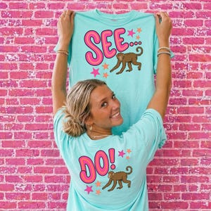 Puede incluir: Camisetas de color azul claro con las palabras "SEE..." y "DO!" en letras rosas con burbujas, cada una con un gráfico de mono marrón y acentos de estrella. Las camisetas se muestran contra una pared de ladrillos rosas.