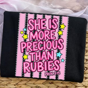 Puede incluir: Camiseta negra con un diseño rosa y blanco. El diseño presenta el texto "SHE IS MORE PRECIOUS THAN RUBIES" en rosa, con acentos de estrellas amarillas y azules. El texto "PROV. 3:15" está en la parte inferior.