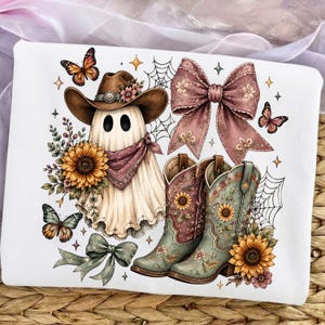May include: A white fabric surface featuring a graphic print of a ghost wearing a brown cowboy hat and a patterned bandana, accompanied by two cowboy boots, sunflowers, butterflies, a large dusty pink bow, a green bow, and spider webs.