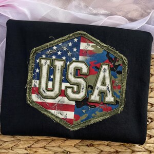 May include: Black t-shirt with a distressed USA patch. The patch is a hexagon shape with a camouflage pattern and the American flag. The word "USA" is in white, block letters.