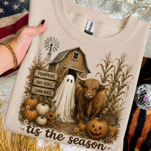 Op de afbeelding: Een zandkleurige sweater met ronde hals en een grafische print van een rustieke schuur, een windmolen, een geest en een Schotse hooglander. Het ontwerp bevat houten borden met PUMPKINS, HAY-RIDES, CORN MAZE en de tekst 'tis the season'.