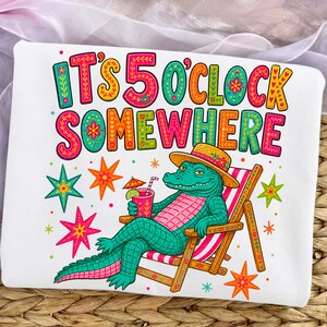 Puede incluir: Camiseta blanca con un gráfico colorido de un caimán relajándose en una silla de playa, sosteniendo un cóctel. El texto "IT'S 5 O'CLOCK SOMEWHERE" se muestra en letras vibrantes y decorativas. El diseño incluye estrellas y una sombrilla de playa.