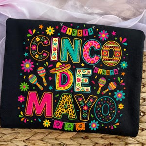 Può includere: Felpa nera con un colorato design del Cinco de Mayo. Il design include le parole "Cinco de Mayo" e "Fiesta" con maracas, fiori e un sombrero. Le lettere hanno vari modelli e colori.