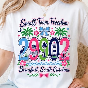 以下が含まれることがあります： Small Town FreedomおよびBeaufort, South Carolinaというテキストが描かれたカラフルなグラフィック付きの白いTシャツ。デザインには数字の29902、青いリボン、ヤシの木、サギ、灯台、錨、ピンクのハイビスカスの花が含まれています。