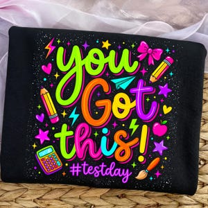 May include: Black t-shirt with a colorful graphic that says "You Got This!" in neon green, orange, and purple. The design includes pencils, stars, a calculator, and the hashtag #testday.
