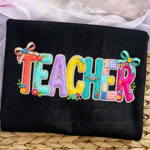 Pode incluir: Camisola preta com a palavra "TEACHER" em letras coloridas aplicadas. As letras são contornadas em branco e decoradas com flores e laços. O desenho está centrado na frente da camisola.