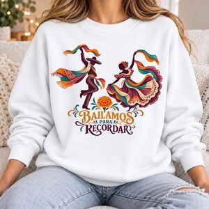 Peut inclure: Un sweat-shirt blanc à col rond orné d'un motif représentant deux danseurs en tenue traditionnelle avec des tissus colorés flottants. Sous les danseurs se trouve une fleur de souci orange et le texte "BAILAMOS PARA RECORDAR" en lettres décoratives.