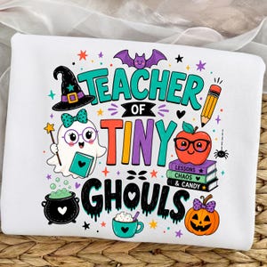 Può includere: Una felpa bianca con una grafica colorata e la scritta "TEACHER OF TINY GHOULS". Il design include un fantasma con occhiali, un cappello da strega, una matita, una mela rossa con occhiali su una pila di libri, un calderone e una zucca.