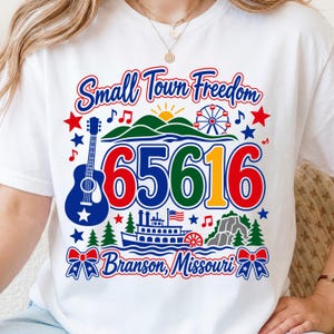 May include: A white t-shirt featuring a graphic with the text Small Town Freedom and Branson, Missouri. The design includes a blue guitar, the number 65616, a ferris wheel, a riverboat, green hills, pine trees, musical notes, and red and blue stars.