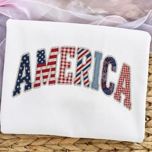 May include: White sweatshirt with the word "AMERICA" in a patchwork design. The letters feature patterns like the American flag, red and white gingham, and a blue denim-like texture, with a fireworks design in the letter "I".