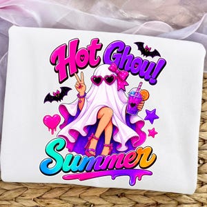 Könnte beinhalten: Weißes T-Shirt mit einem Cartoon-Geist mit Sonnenbrille und Schleife, der ein Getränk hält und das Friedenszeichen zeigt. Die Worte "Hot Ghoul!" und "Summer" sind in farbenfrohem, stilisiertem Text geschrieben, zusammen mit Fledermäusen, Herzen und Sternen.