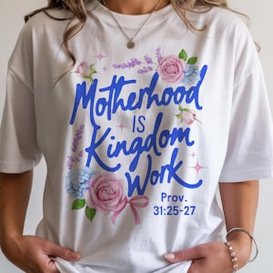 May include: White t-shirt with the text "Motherhood Is Kingdom Work" in blue script, surrounded by watercolor flowers in pink, blue, and purple. The shirt also includes the Bible verse Proverbs 31:25-27.