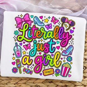 Pode incluir: Camisola branca com um gráfico colorido que diz "Literally just a girl". O design inclui um laço rosa, um sapato de cristal, borboletas, estrelas, corações e outras ilustrações divertidas.