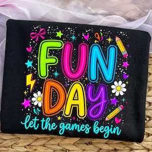 Op de afbeelding: Zwart T-shirt met een kleurrijk "FUN DAY" ontwerp. Het ontwerp bevat de tekst "let the games begin", sterren, harten, potloden en bloemen. De letters zijn omlijnd in heldere kleuren.