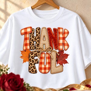 Peut inclure: Un t-shirt blanc à manches courtes arborant le mot THANKFUL dans un graphisme à motifs variés. Les lettres alternent entre un motif à carreaux orange et blanc, un imprimé léopard et une texture de toile de jute, avec un nœud en ficelle.