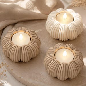 Könnte beinhalten: Drei kürbisförmige Kerzenhalter in Creme- und Beigetönen. Jeder Halter hat eine gerippte, vertikale Textur und eine kreisförmige Öffnung in der Mitte, die ein kleines, rundes, weißes Teelicht mit einer warm leuchtenden Flamme enthält.