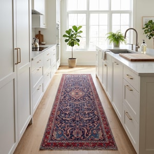 May include: A long, rectangular rug with an intricate floral pattern in shades of navy blue, red, and cream. The rug is placed in a kitchen with white cabinets and light wood flooring. The rug's design features a central medallion and detailed borders.